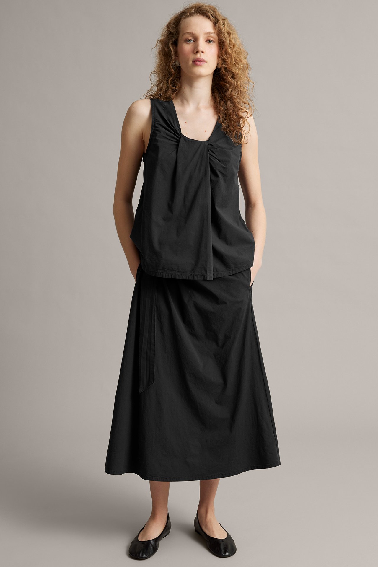 Frontvy: kvinna i svart linne i poplin med omlottringning, svart midikjol och svarta ballerinaskor.