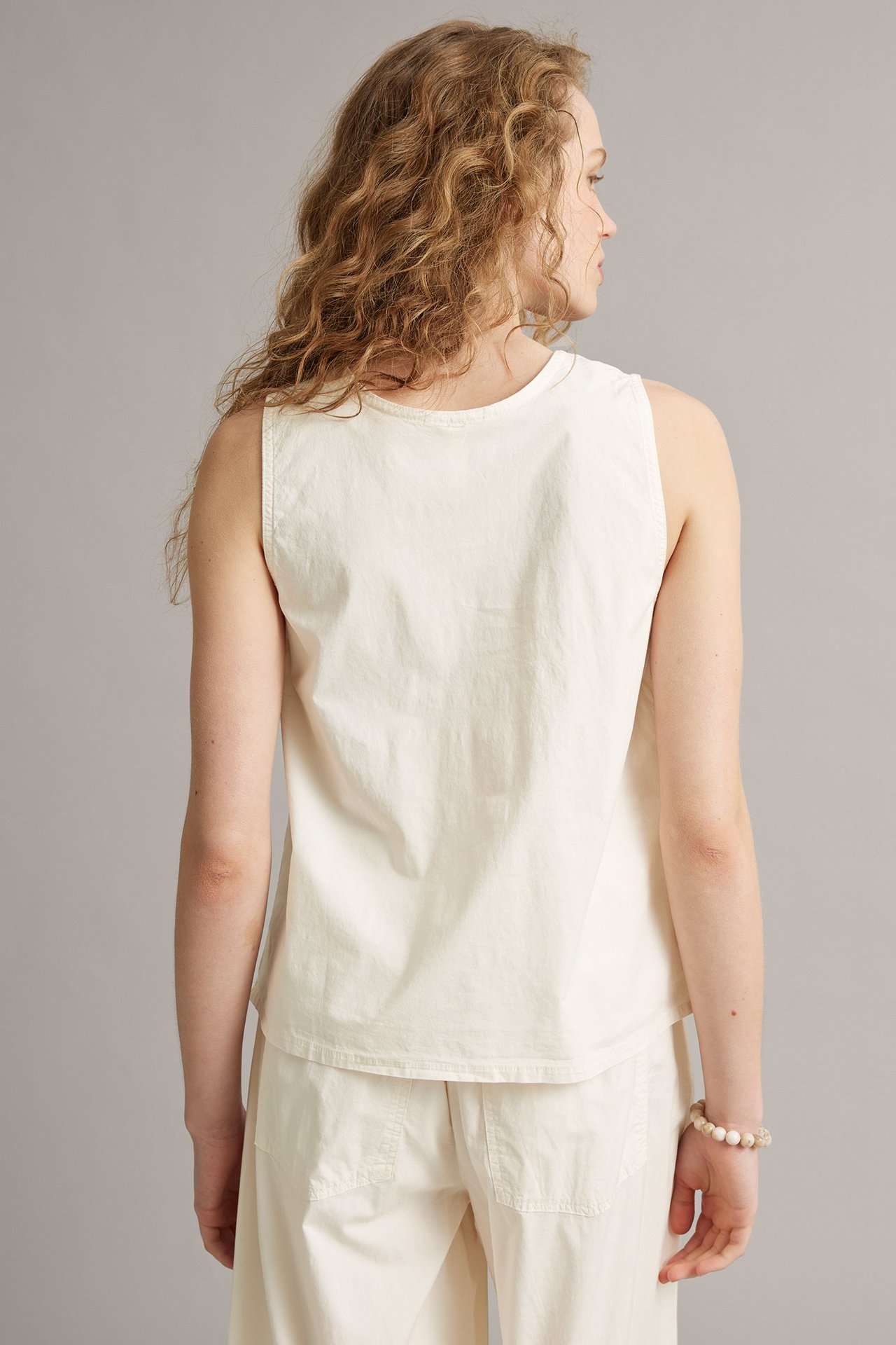 Naisen taka-sivukuva: valkoinen kudottu toppi, valkoiset leveät housut ja vaalea helmikoristeltu rannekoru.