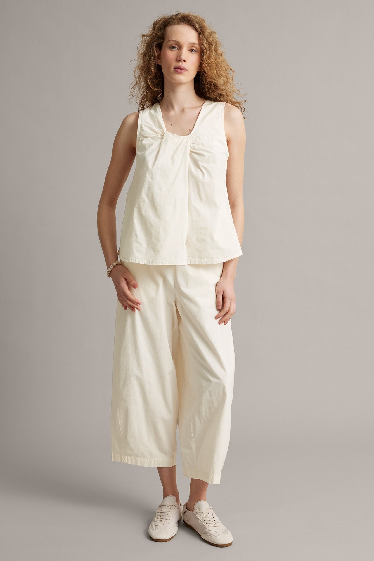 Frontutsikt: Kvinne i hvit ermeløs topp med samlet utringning, capribukser og hvite joggesko.
