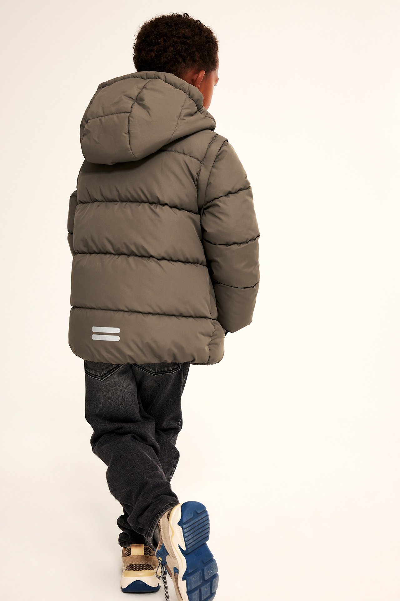 Bakfra av et barn i en khakifarget pufferjakke med hette, mørkegrå jeans og flerfargede joggesko.