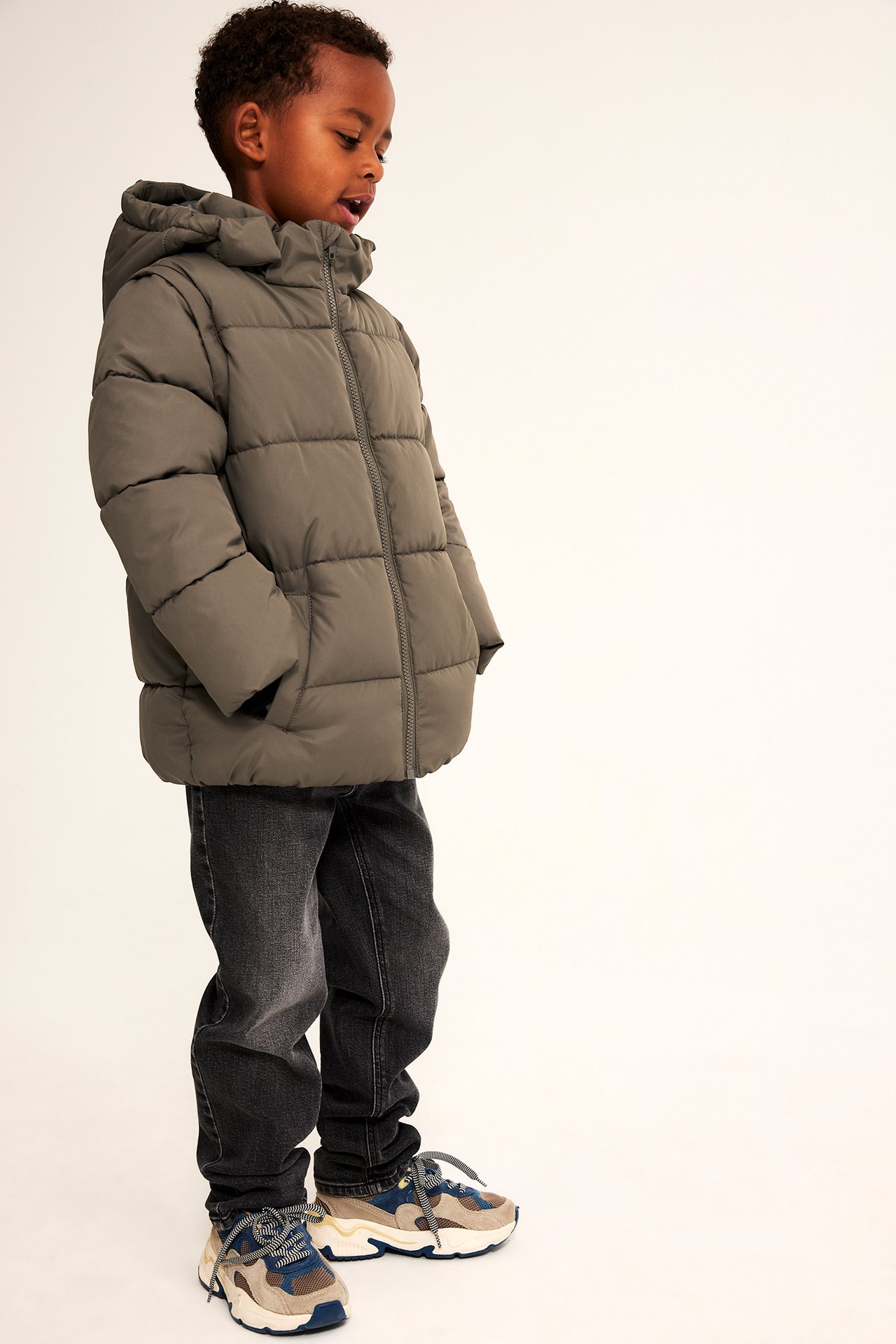 Sidovy av ett barn i grön puffig jacka med huva, svarta jeans och flerfärgade sneakers.