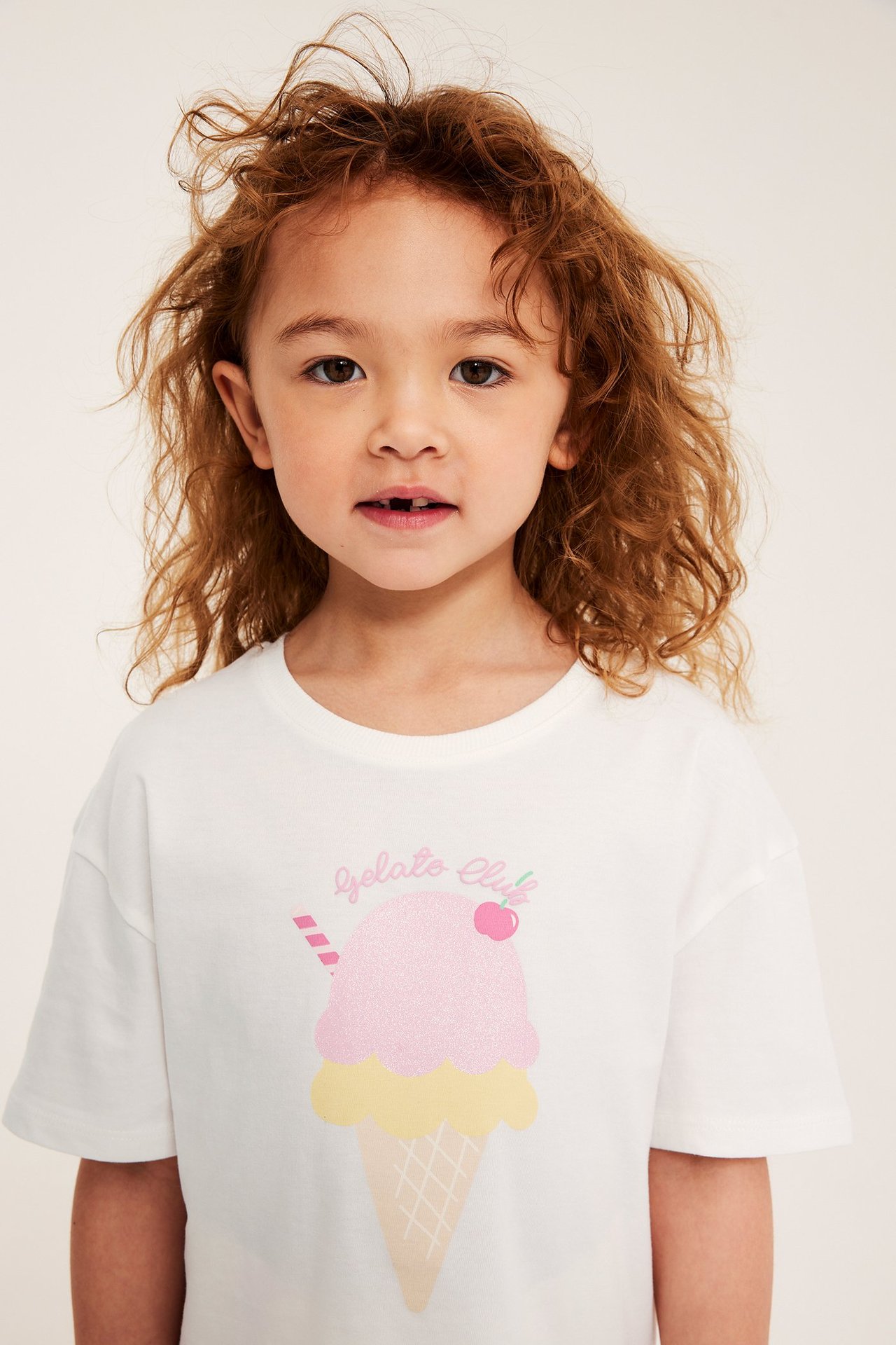Lähikuva edestä lapsesta valkoisessa puuvilla-T-paidassa, jossa on kimalteleva jäätelökuvio.
