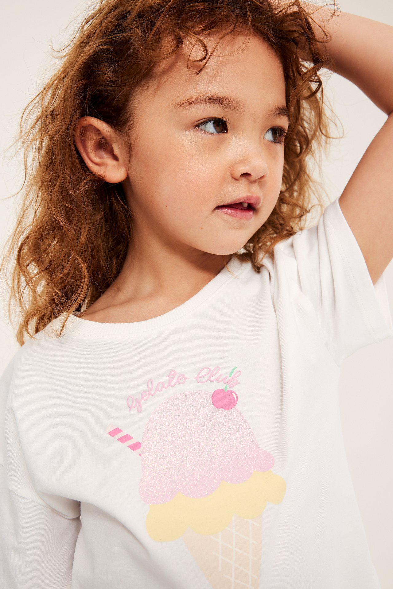 Närbild: Ett barn bär en vit bomulls-t-shirt med glittrigt glasstryck och "Gelato Club".
