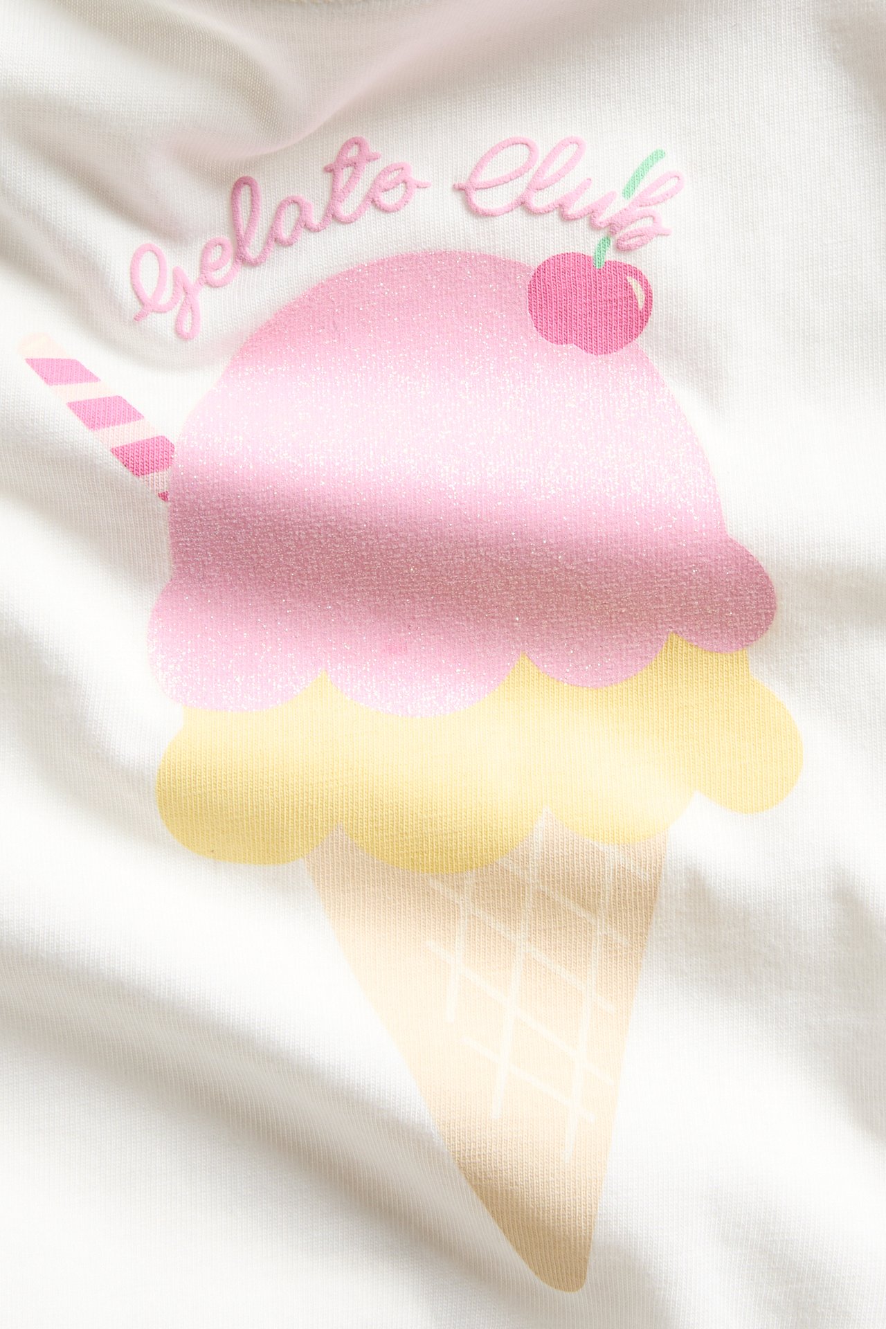 Lähikuva valkoisesta puuvillaisesta T-paidasta, jossa vaaleanpunainen ja keltainen kimalteleva jäätelöprintti.