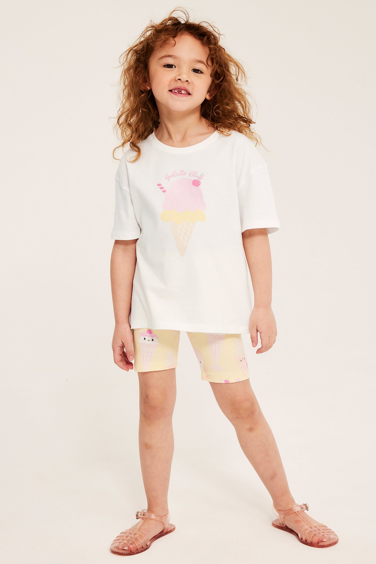 Framifrån: Barn i vit bomulls-T-shirt med glittrigt glasstryck, gula mönstrade shorts och rosa gelésandaler.