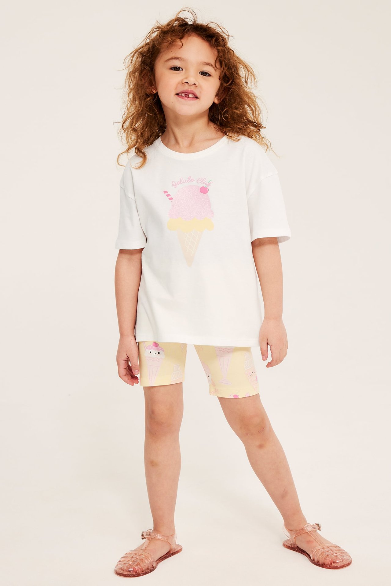 Widok z przodu: Dziecko w białym bawełnianym T-shircie z brokatowym nadrukiem loda, żółtych wzorzystych szortach i różowych sandałach.
