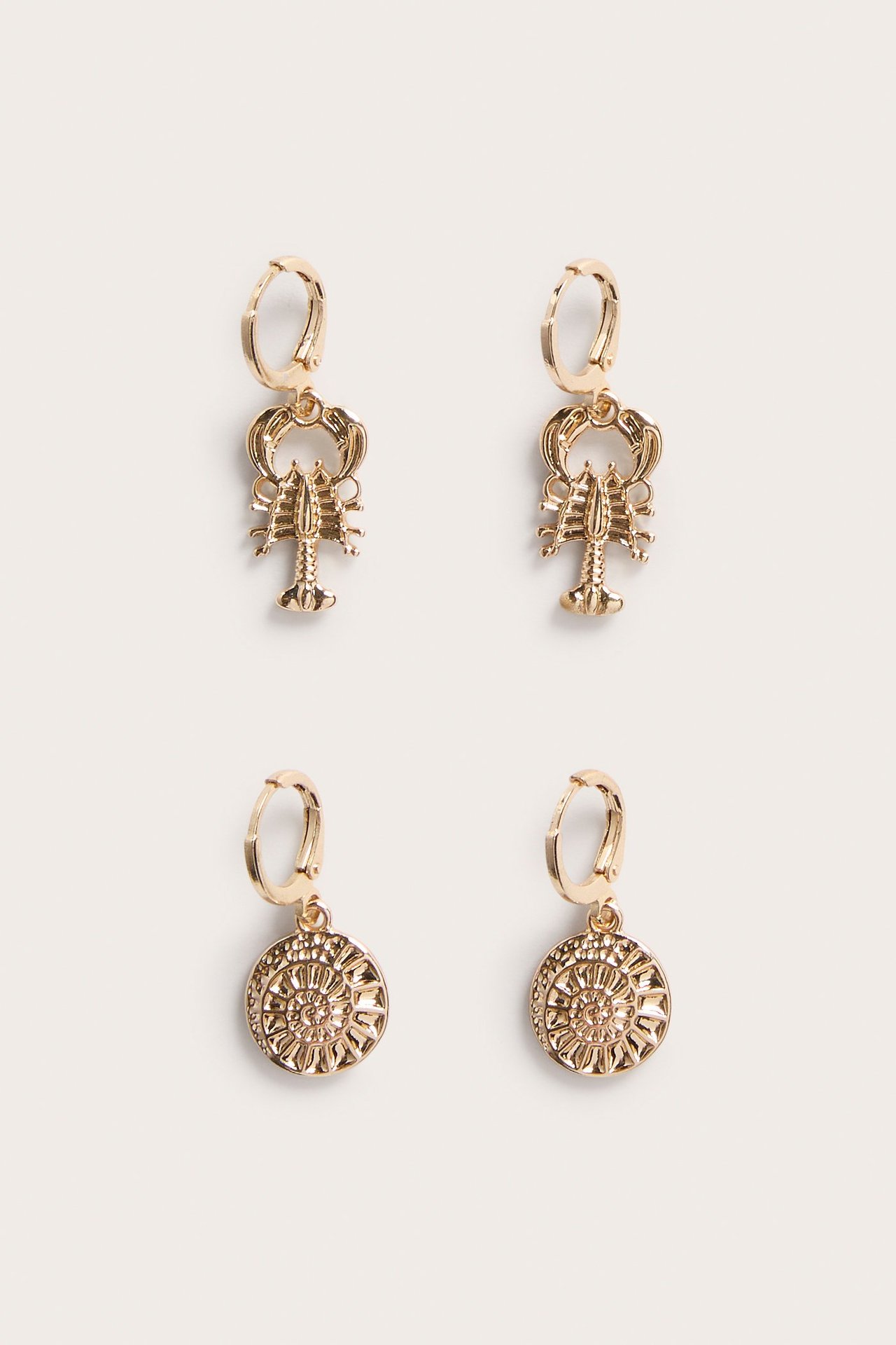 Lähikuva kullansävyisistä metallikorvakoruista; yhdet hummerikorulla, toiset kuvioidulla medaljongilla.