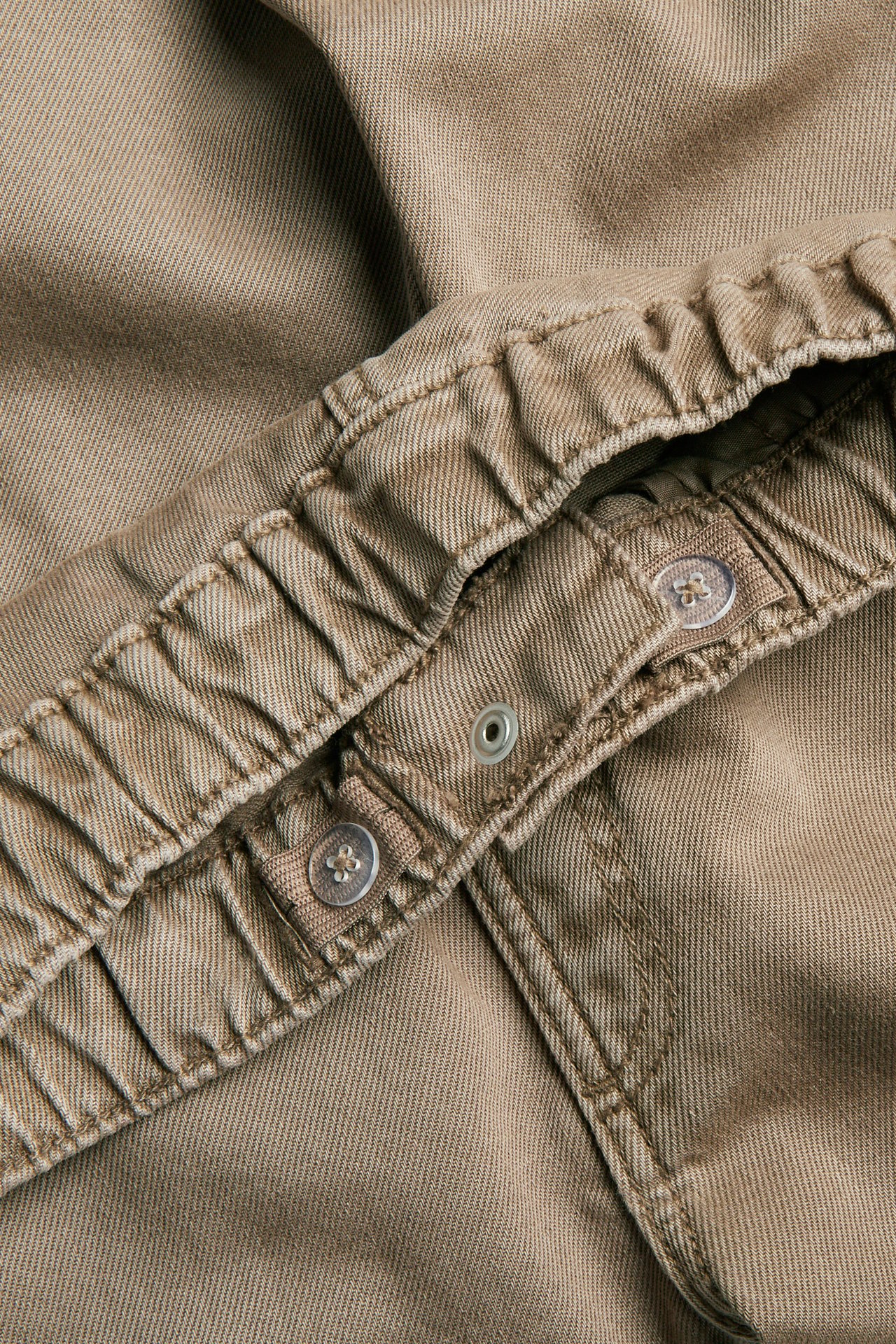 Närbild på beiga denimbyxors midjeband med resår, justerbara knappar och tryckknapp.