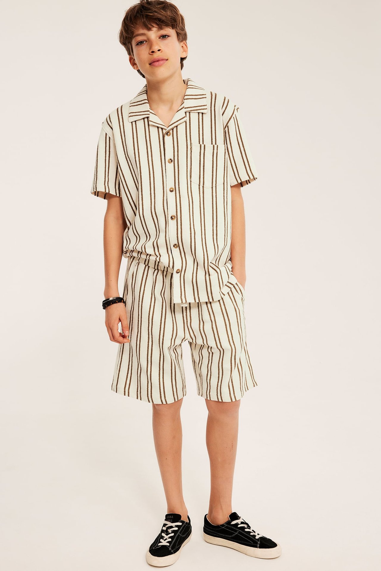 Barn bär en beige randig bomullsskjorta och matchande shorts, svarta sneakers och ett mörkt armband.