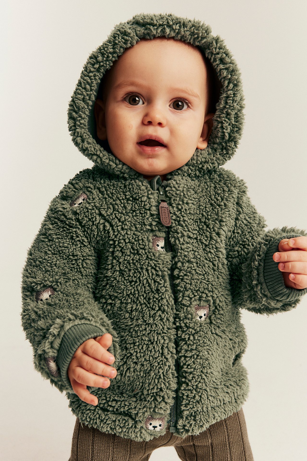 Framifrån, baby bär mörkgrön lurvig huvjacka med igelkottbroderi och bruna ribbade leggings.