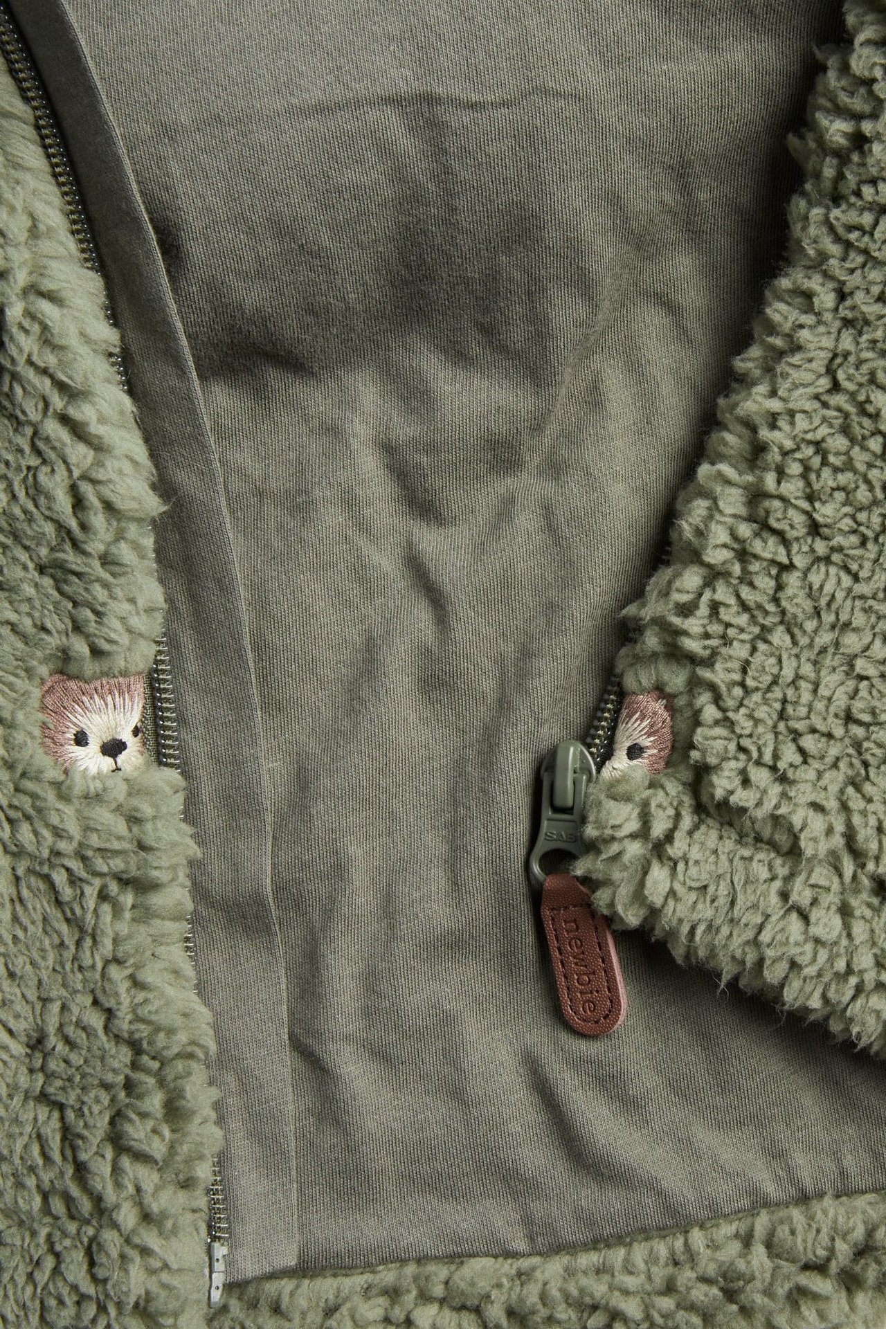 Lähikuva vihreästä teddy-takista, jossa vetoketju ja brodeeratut karhun kasvot.