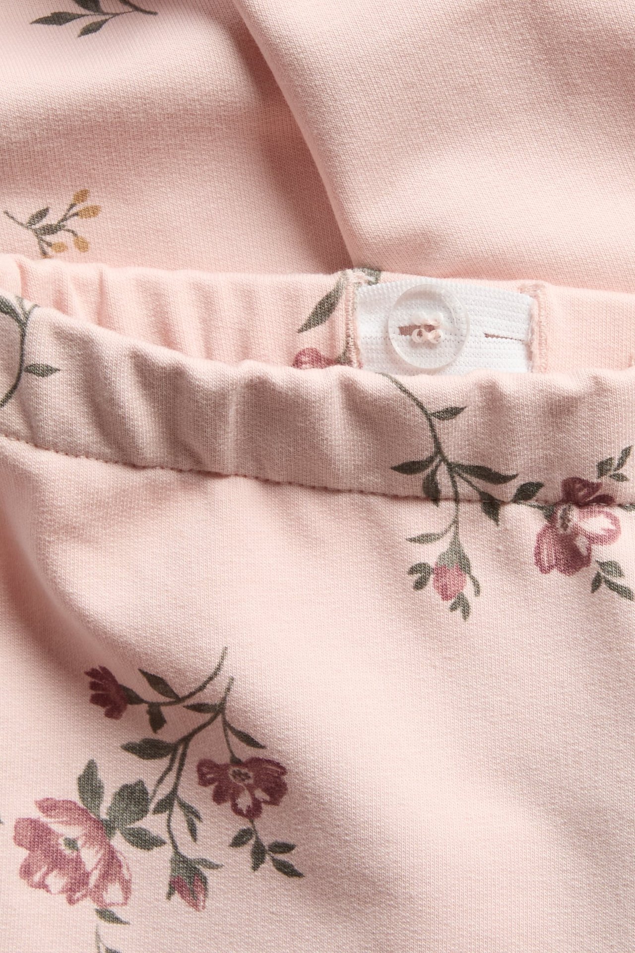Nærbilde av lys rosa blomstrete jerseybukser med justerbar elastisk linning og hvit knapp.