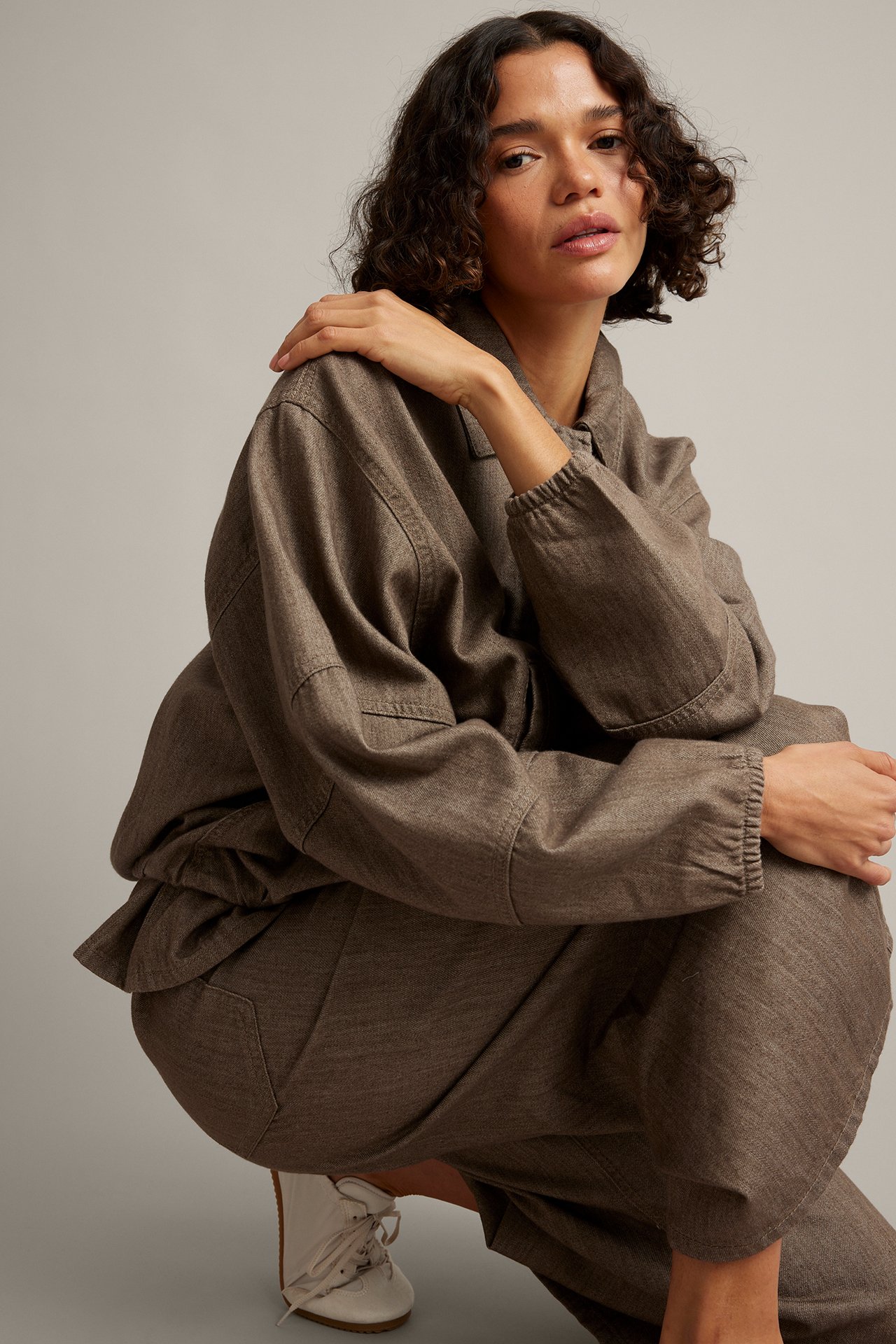Frontvy: Kvinnlig modell i oversized brun-grå texturerad skjorta och matchande lösa byxor, med gräddvita skor.
