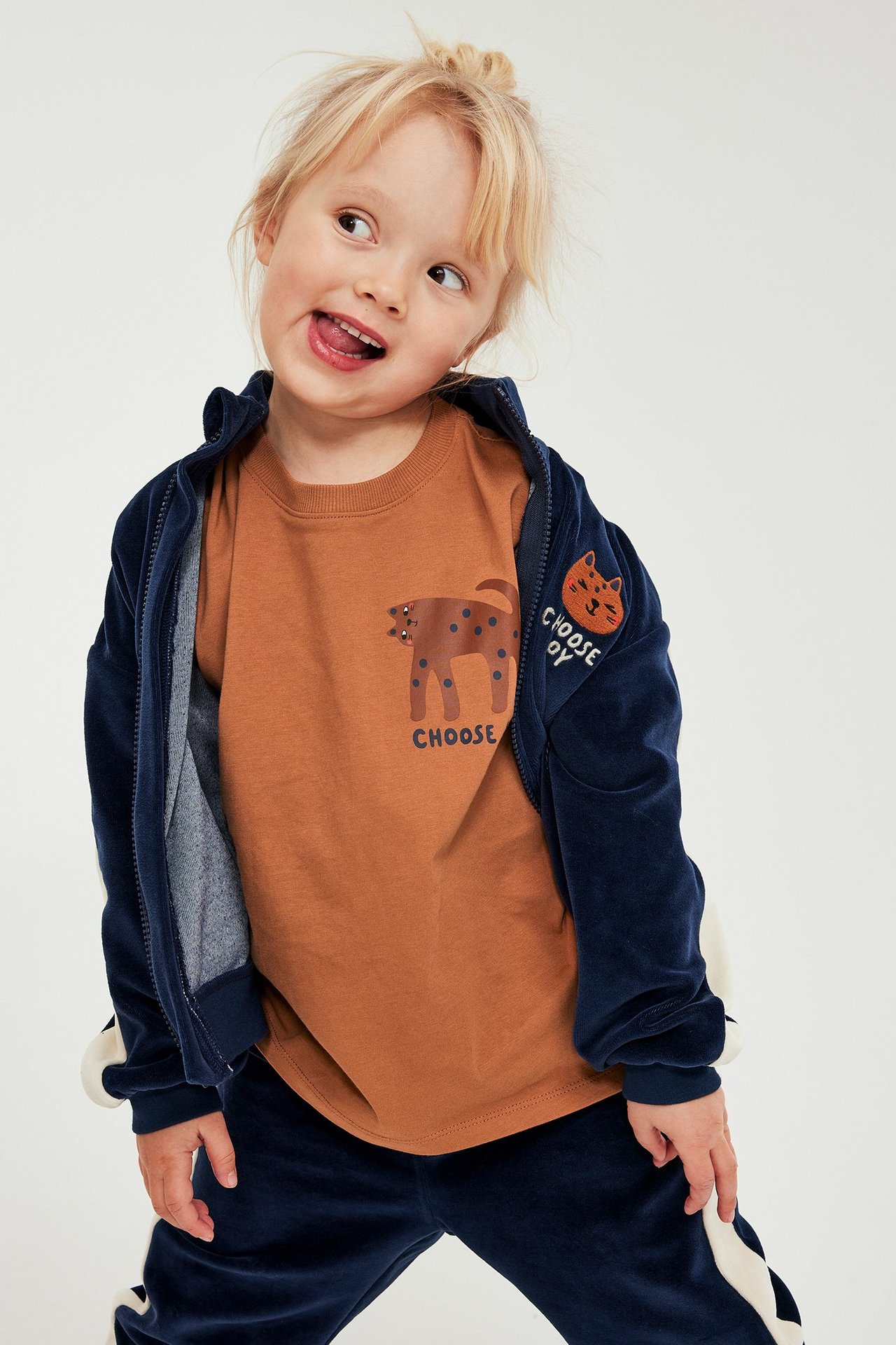 Framifrån ses ett barn i en brun kortärmad t-shirt med djurtryck och mörkblå velourträningsdräkt.