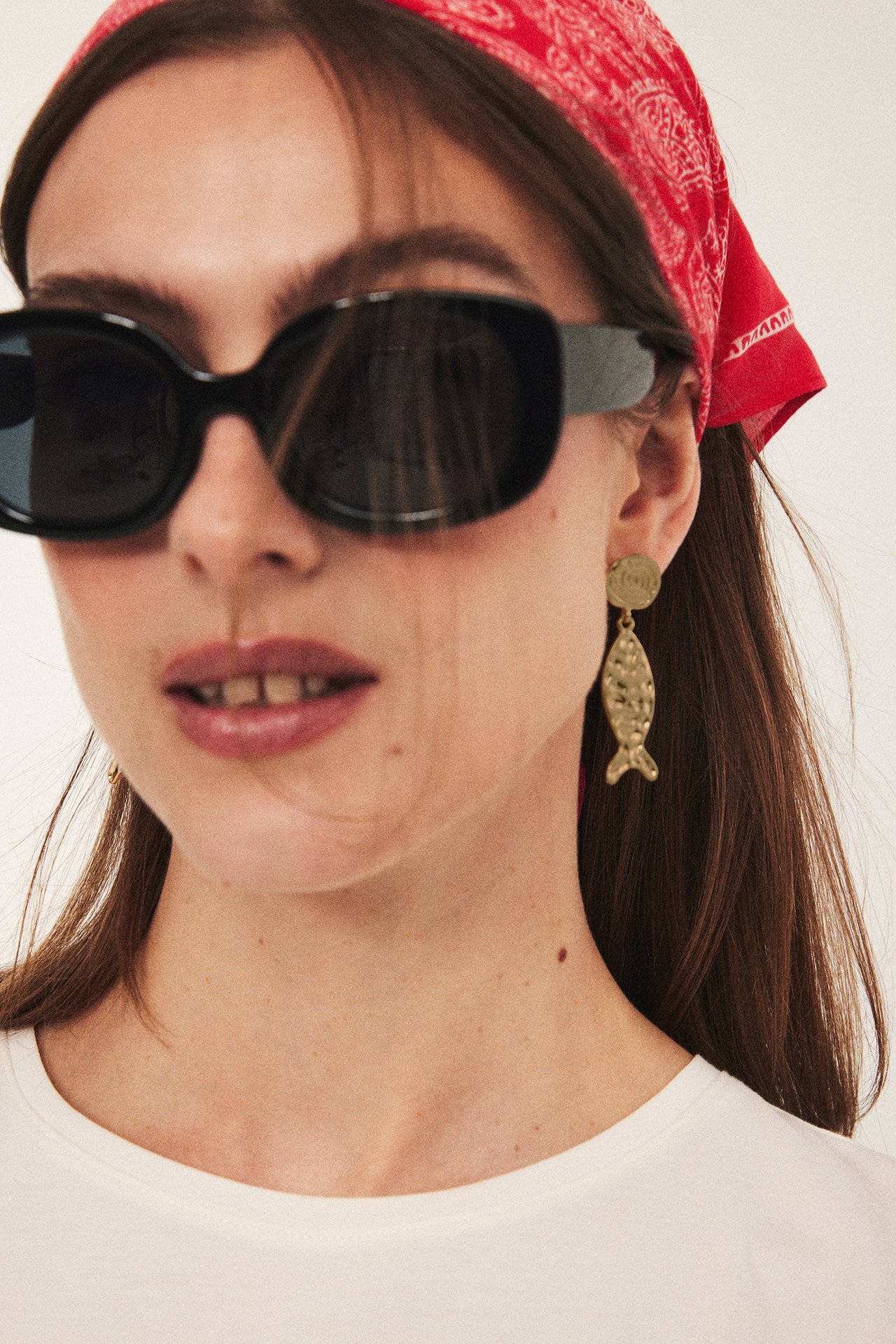 Lähikuva naisesta, jolla on kultaiset kalanmuotoiset metallikorvakorut, mustat aurinkolasit ja punainen bandana.