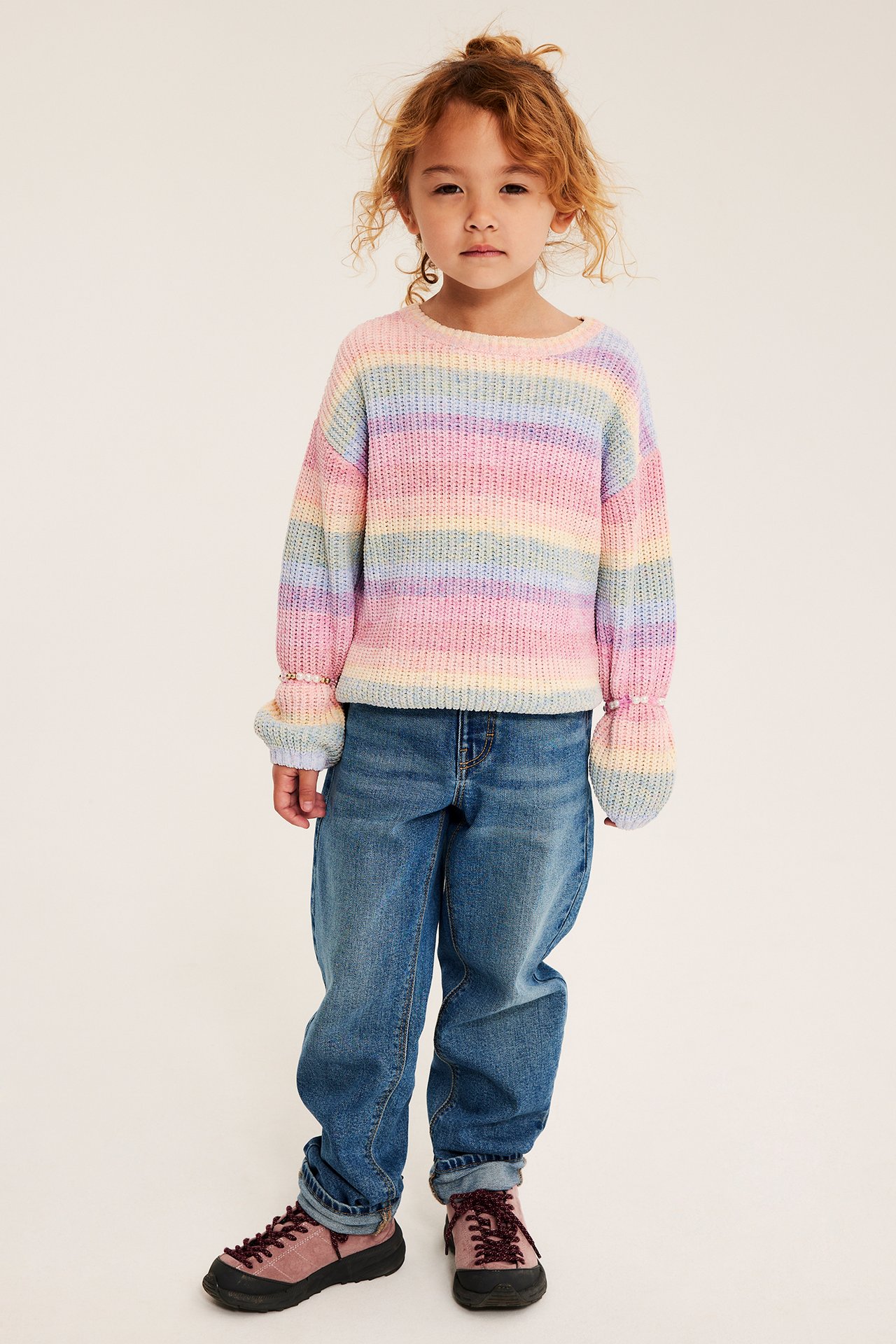 Barns framifrån: en pastellrandig stickad tröja, blå denimjeans och mörkrosa snörskor.