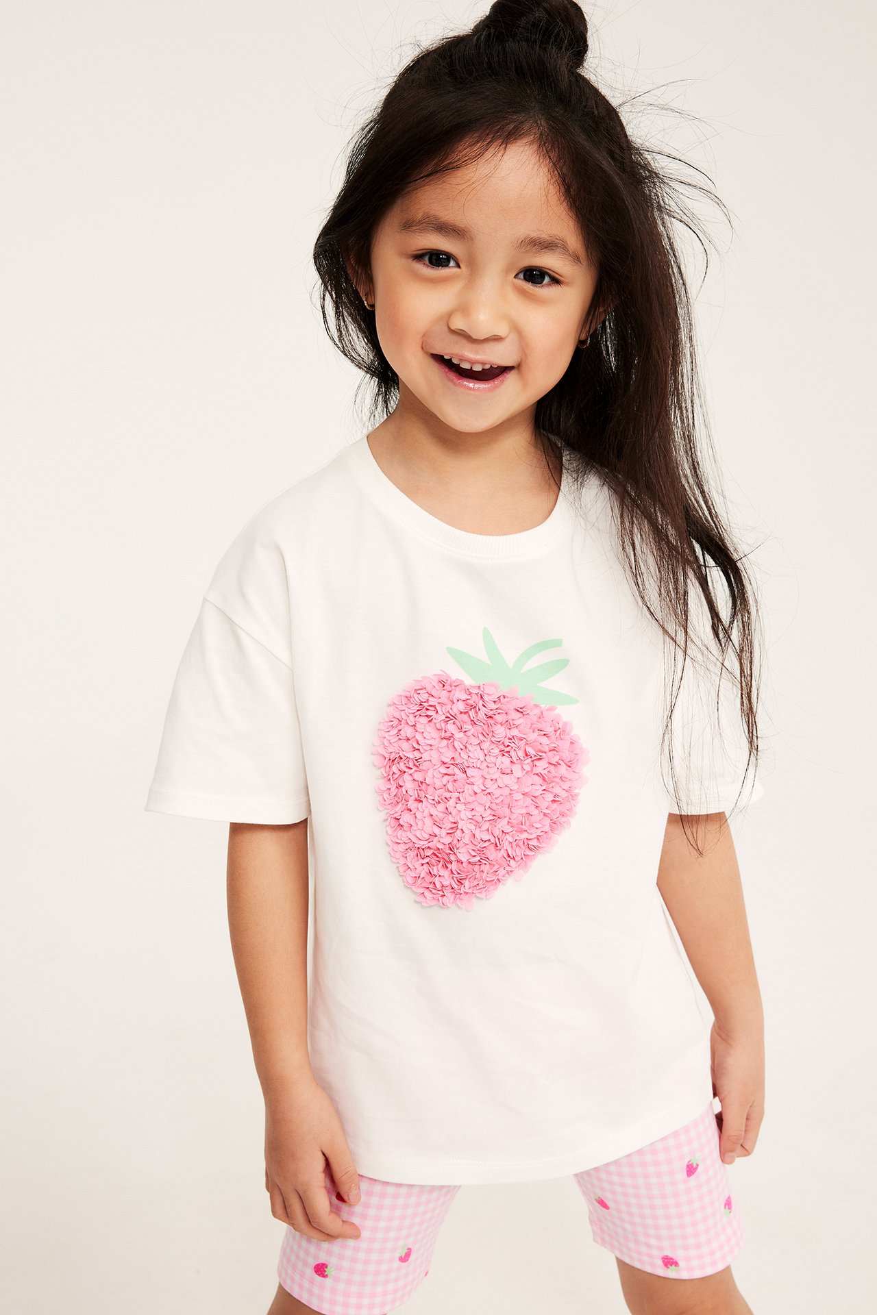 Ett barn i naturvit bomulls-T-shirt med rosa 3D-jordgubbe i mesh, plus rosa rutiga shorts.