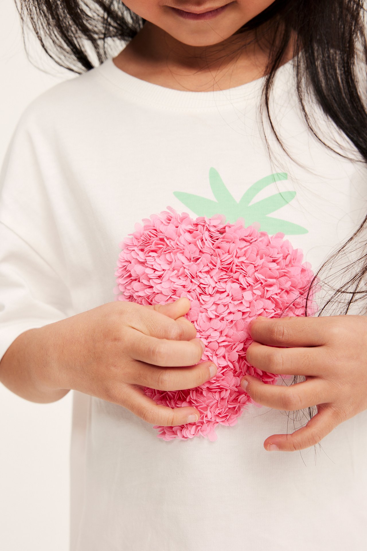 Närbild på ett barn som bär en offwhite bomullst-shirt med rosa nätjordgubbsapplikation, händerna rör vid den.