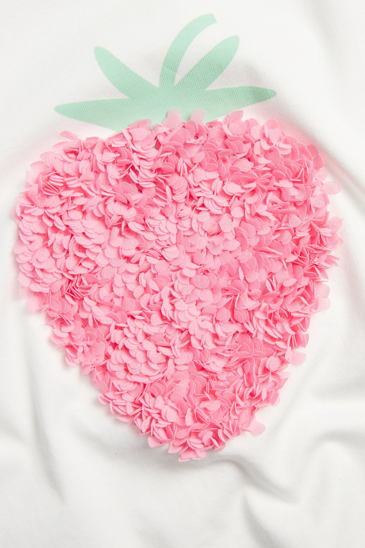 Närbild av offwhite bomulls-t-shirt med rosa 3D mesh-jordgubbsapplikation och gröna tryckta blad.