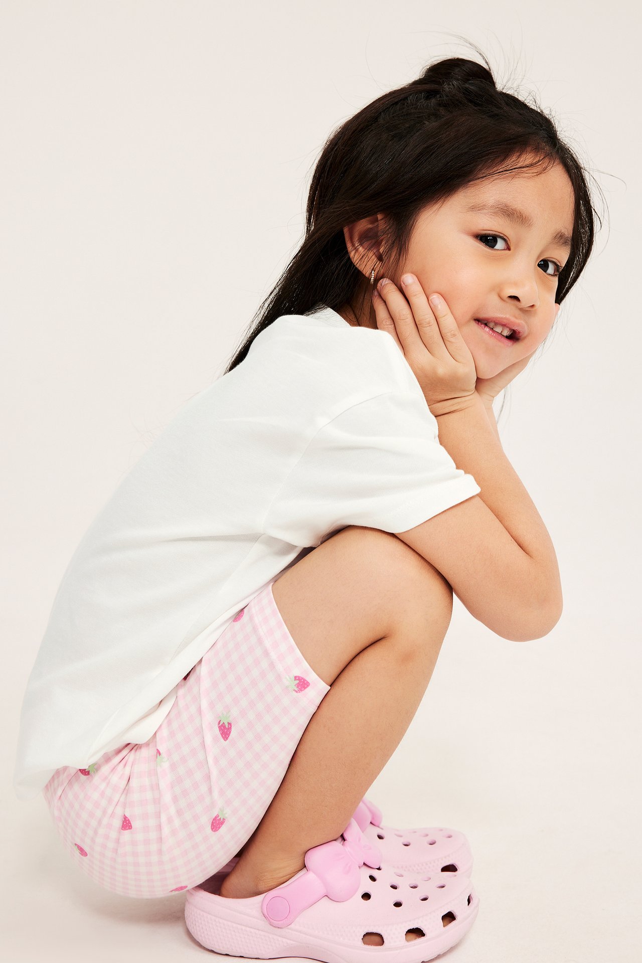Framifrån, ett barn i en benvit T-shirt, rosa rutiga shorts med jordgubbsmönster och rosa tofflor.