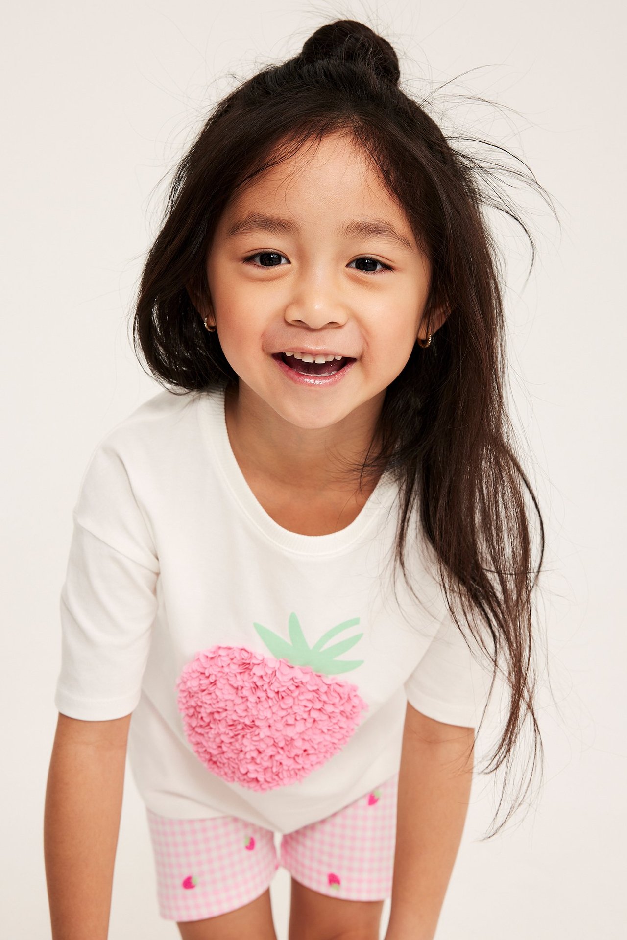 Frontvy av ett barn i en offwhite bomullst-shirt med rosa jordgubbsapplikation och rosa rutiga shorts.