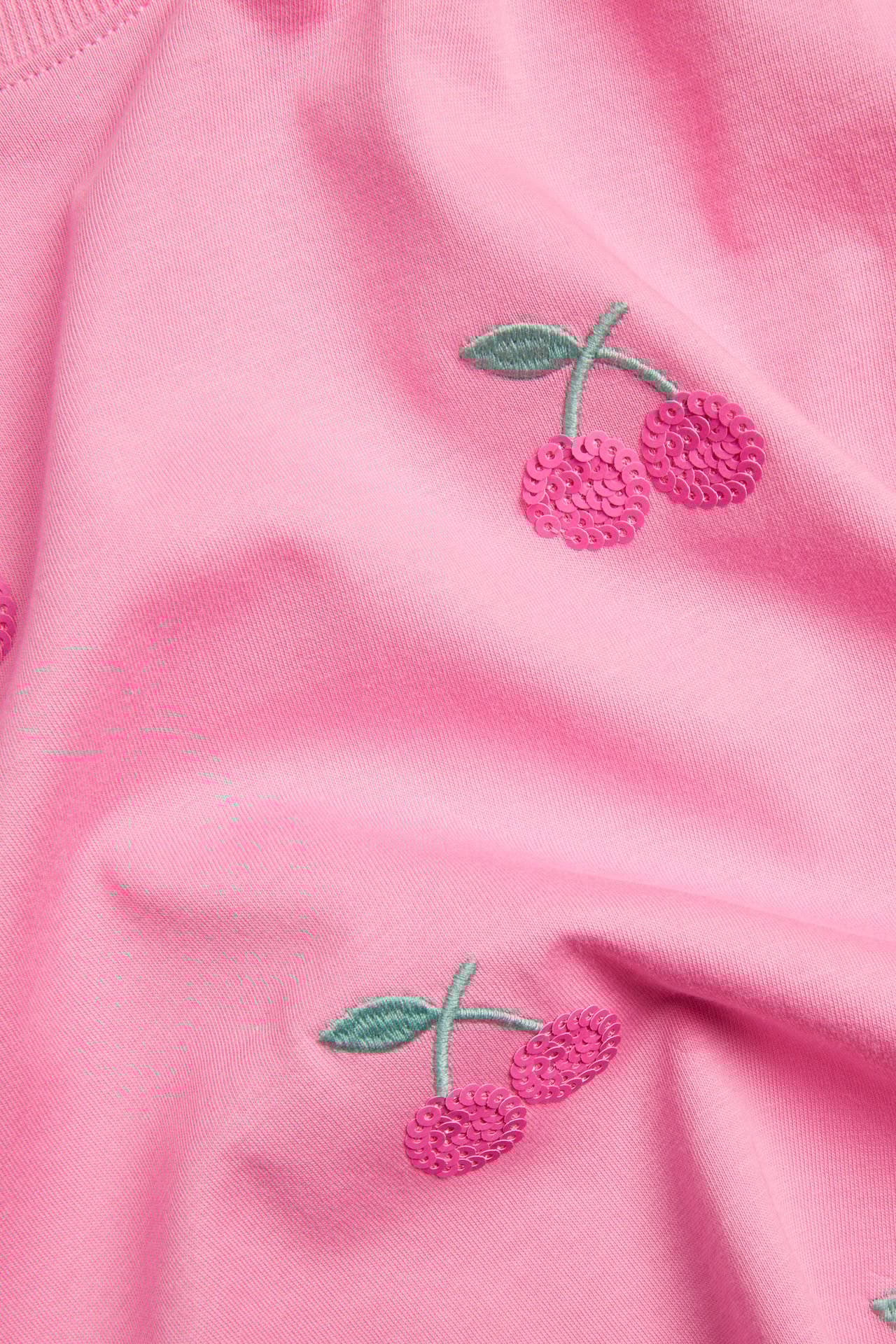 Närbild på en rosa bomullströja med glittriga rosa körsbärspaljetter och gröna stjälkar.