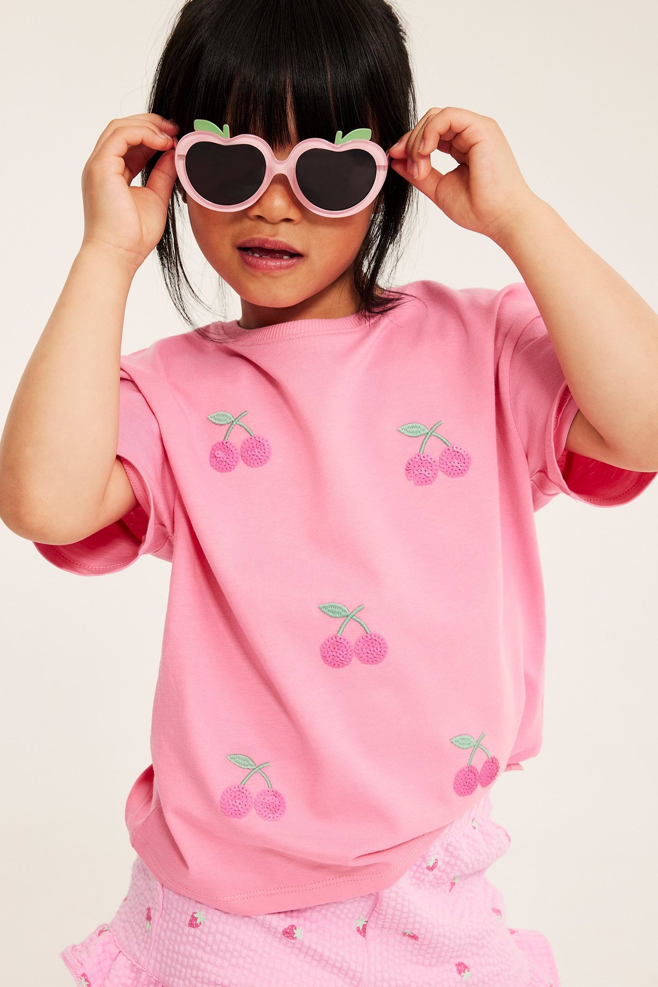 Frontbilde: Barn i rosa kortermet t-skjorte med paljettkirsebær, rosa shorts og rosa hjerteformede solbriller.