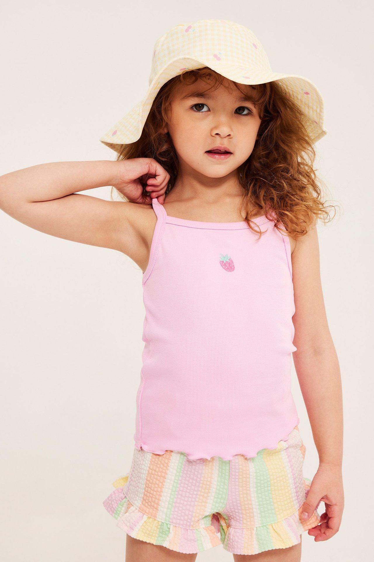 Widok z przodu: Dziecko w jasnoróżowym prążkowanym topie z haftem truskawki, wielobarwnych marszczonych szortach i kapeluszu bucket.