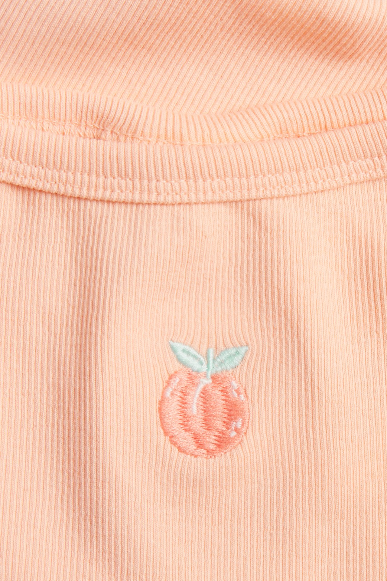 Lähikuva edestä, korallinvärisestä, ribatusta lasten topista pienellä kirjotulla persikalla.