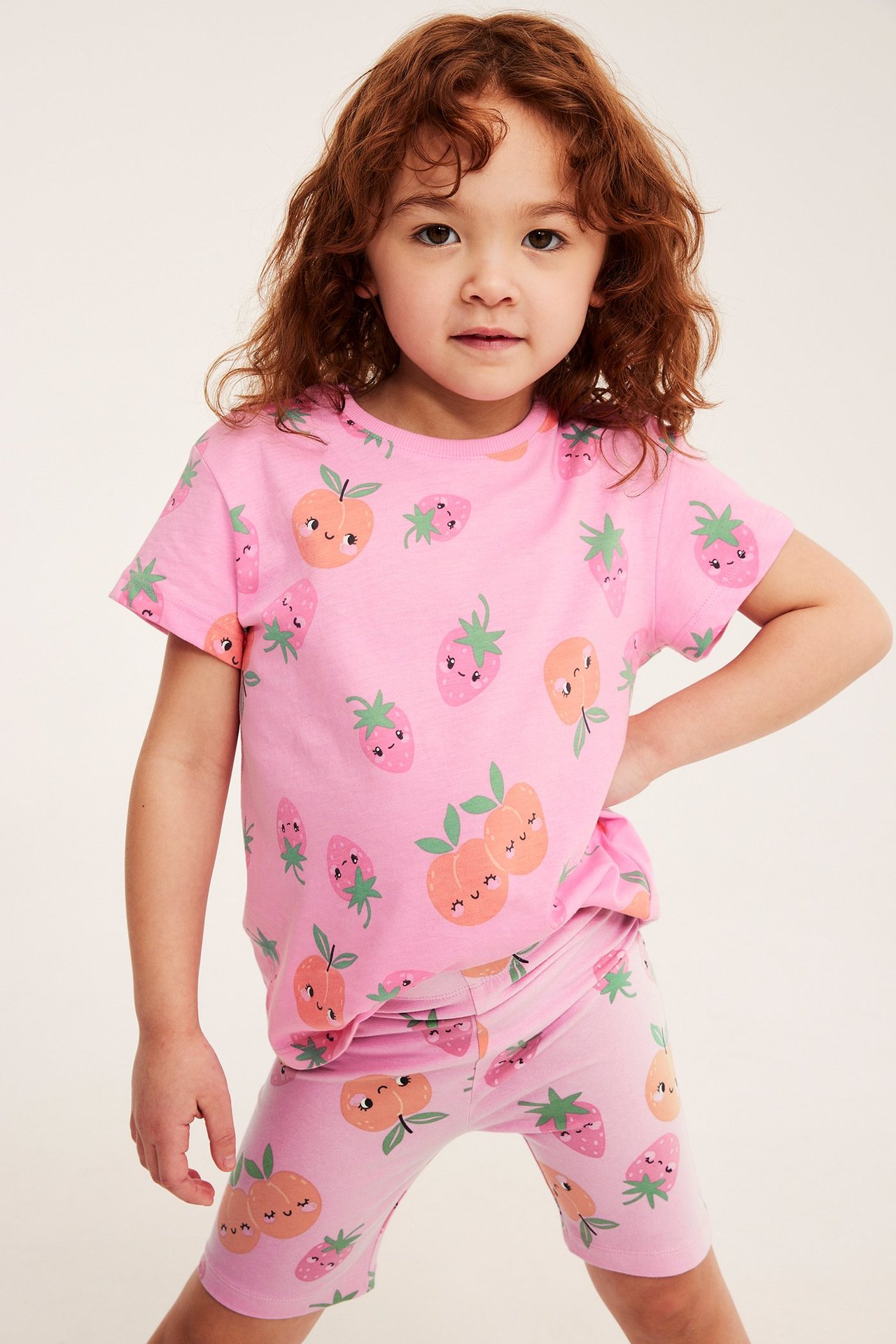 Edestä, lapsi yllään vaaleanpunaiset puuvillatrikoiset hedelmäkuvioiset pyöräilyshortsit ja yhteensopiva t-paita.