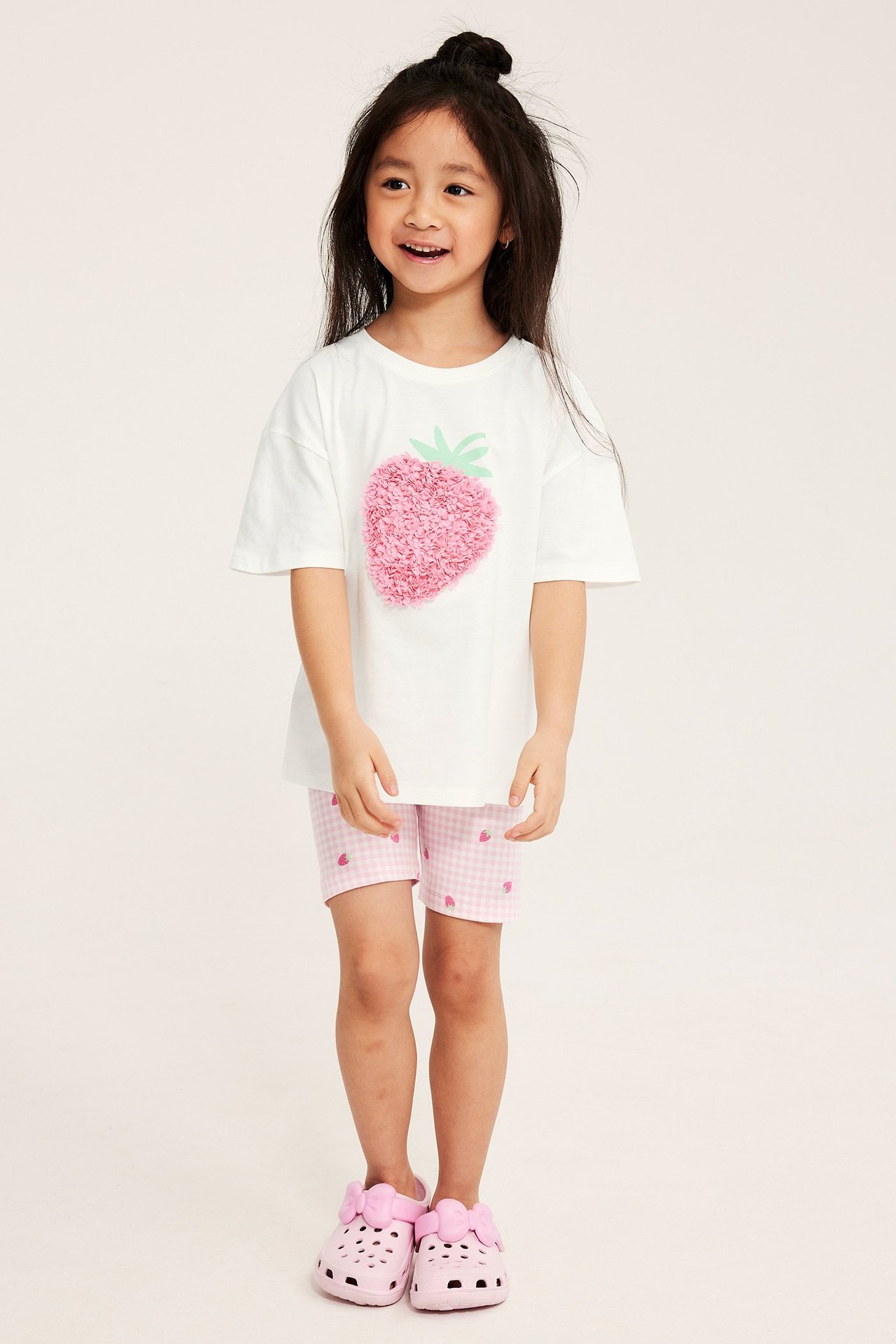 Framifrån: Barn bär rosa rutiga jordgubbs-cykelbyxor, vit jordgubbs-t-shirt och rosa träskor.