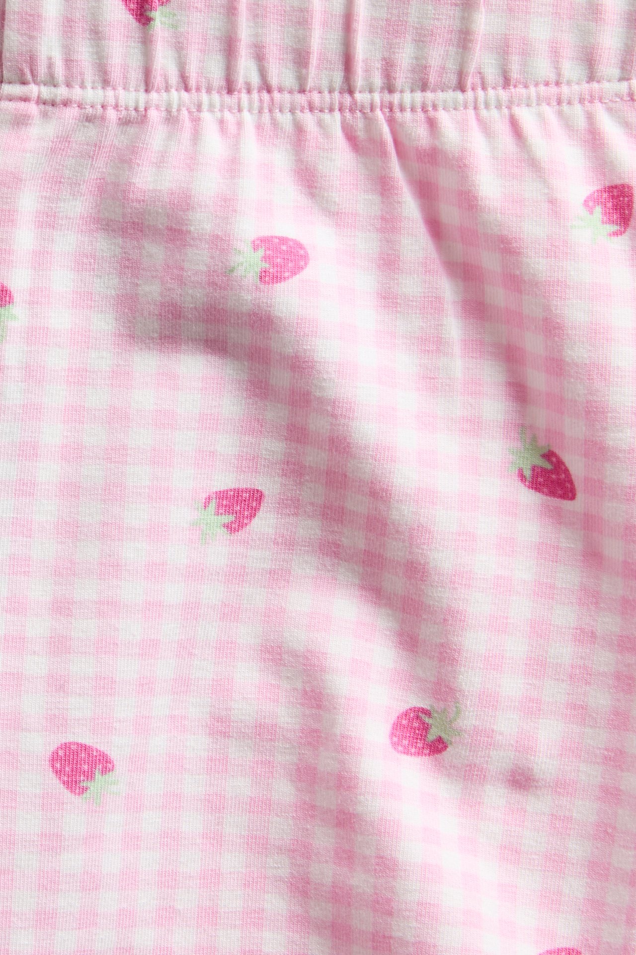 Lähikuva vaaleanpunaisista ja luonnonvalkoisista ruudullisista puuvillatrikooshortseista mansikkakuviolla.