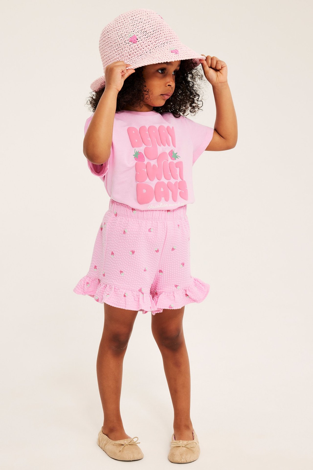 Barn bär rosa bomullst-shirt med text, matchande jordgubbsshorts, rosa stråhatt, beige skor.
