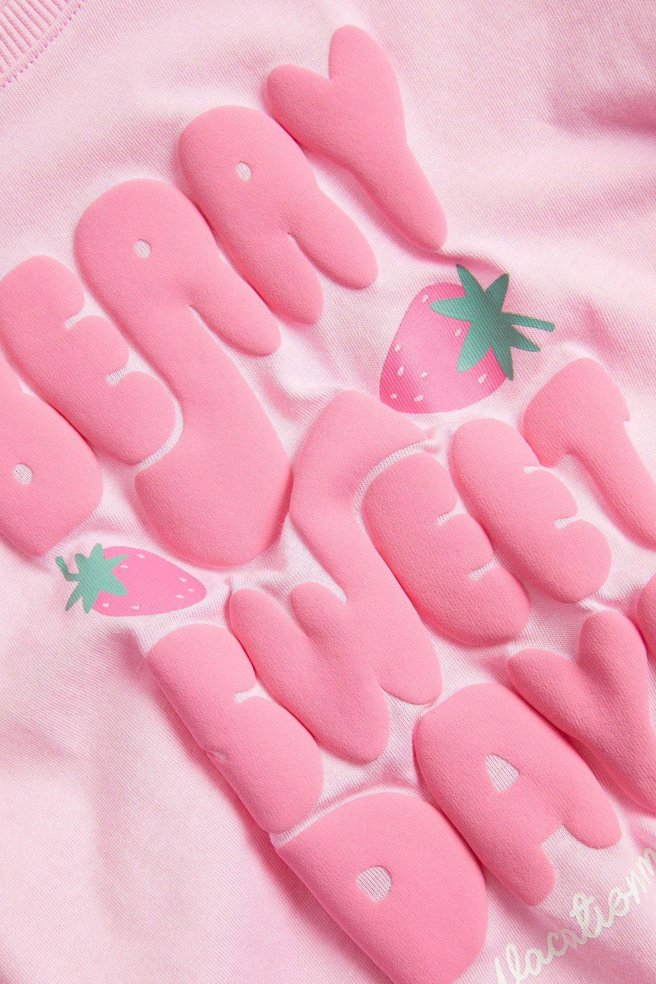 Närbild på en ljusrosa t-shirt i bomull för barn med upphöjt rosa text- och jordgubbstryck.