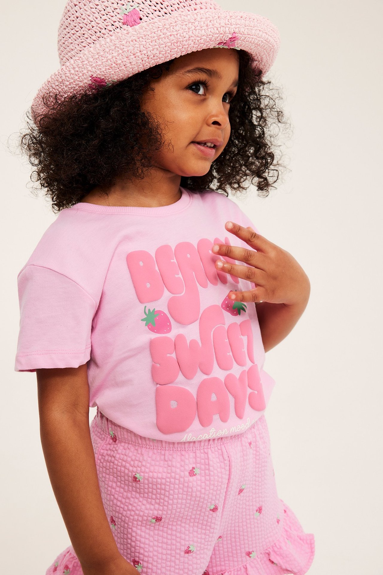 Frontvy: Ett barn i ljusrosa bomulls-t-shirt med text- och jordgubbstryck, rosa hatt och shorts.