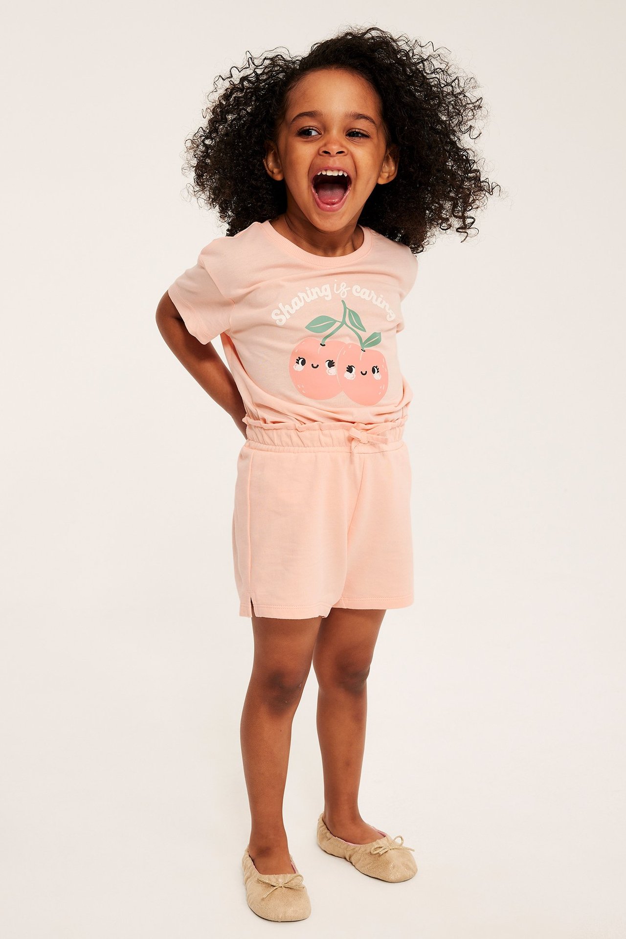 Edestäpäin: lapsi korallinvärisessä puuvilla t-paidassa persikka-printillä, yhteensopivissa shortseissa, beigenväriset ballerinat.