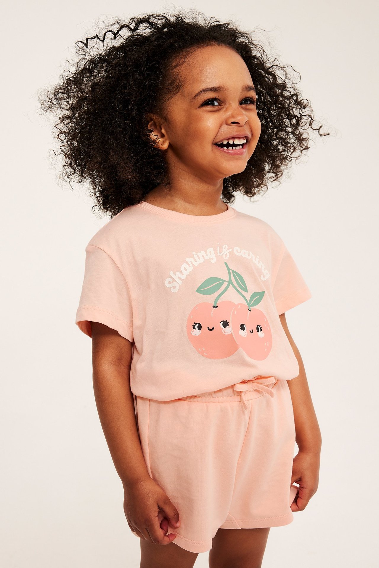 Frontvy av ett barn i en korallfärgad bomulls-t-shirt med persikoprint, stylad med matchande korallfärgade shorts.
