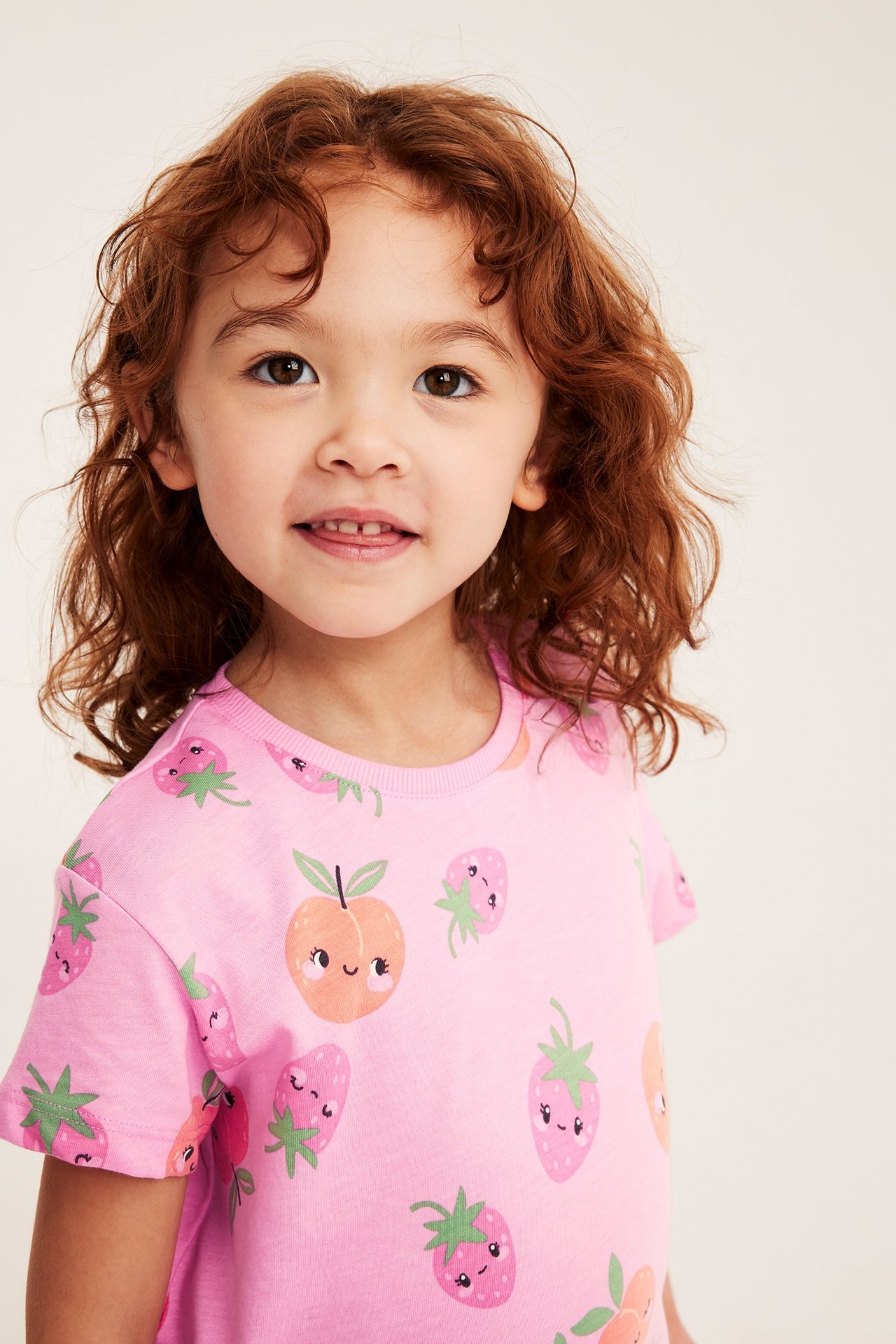 Närbild, framifrån, av ett barn som bär en rosa bomulls-T-shirt med jordgubbs- och persikomönster.
