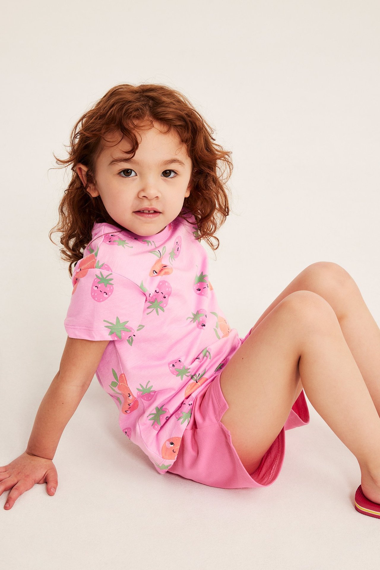 Widok z przodu: dziecko w różowym bawełnianym T-shircie w truskawki i brzoskwinie, plus różowe szorty.