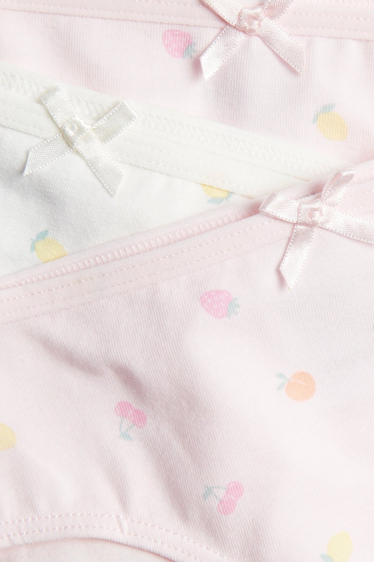 Lähikuva kolmesta lapsen alushousuparista: kaksi vaaleanpunaista ja yksi valkoinen, hedelmäkuviolla ja pienillä satiininauhoilla.