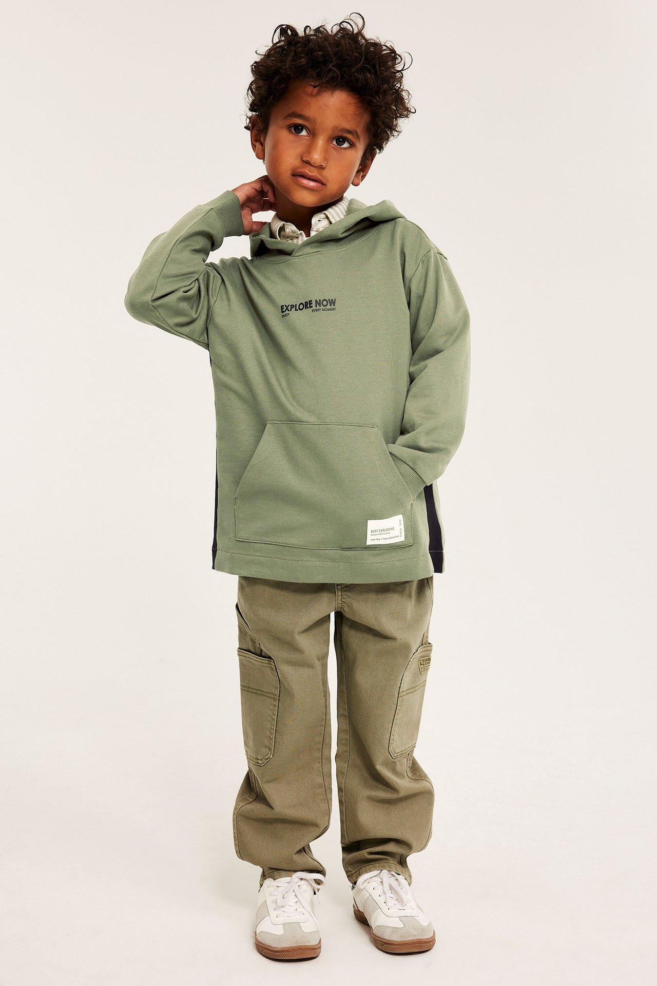 Edestä: poika tummanvihreässä puuvillahupparissa, oliivinruskeissa rahtihousuissa ja valkoisissa tennareissa.