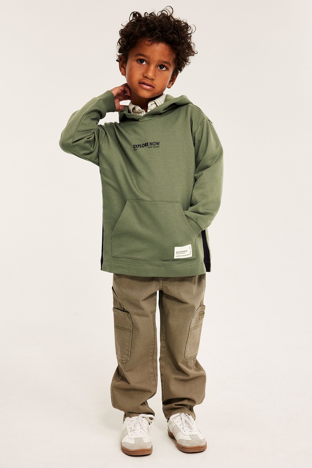 Edestä näkymä lapsesta tummanvihreässä puuvillahupparissa tekstillä, ruskeissa cargohousuissa ja valkoisissa lenkkareissa.