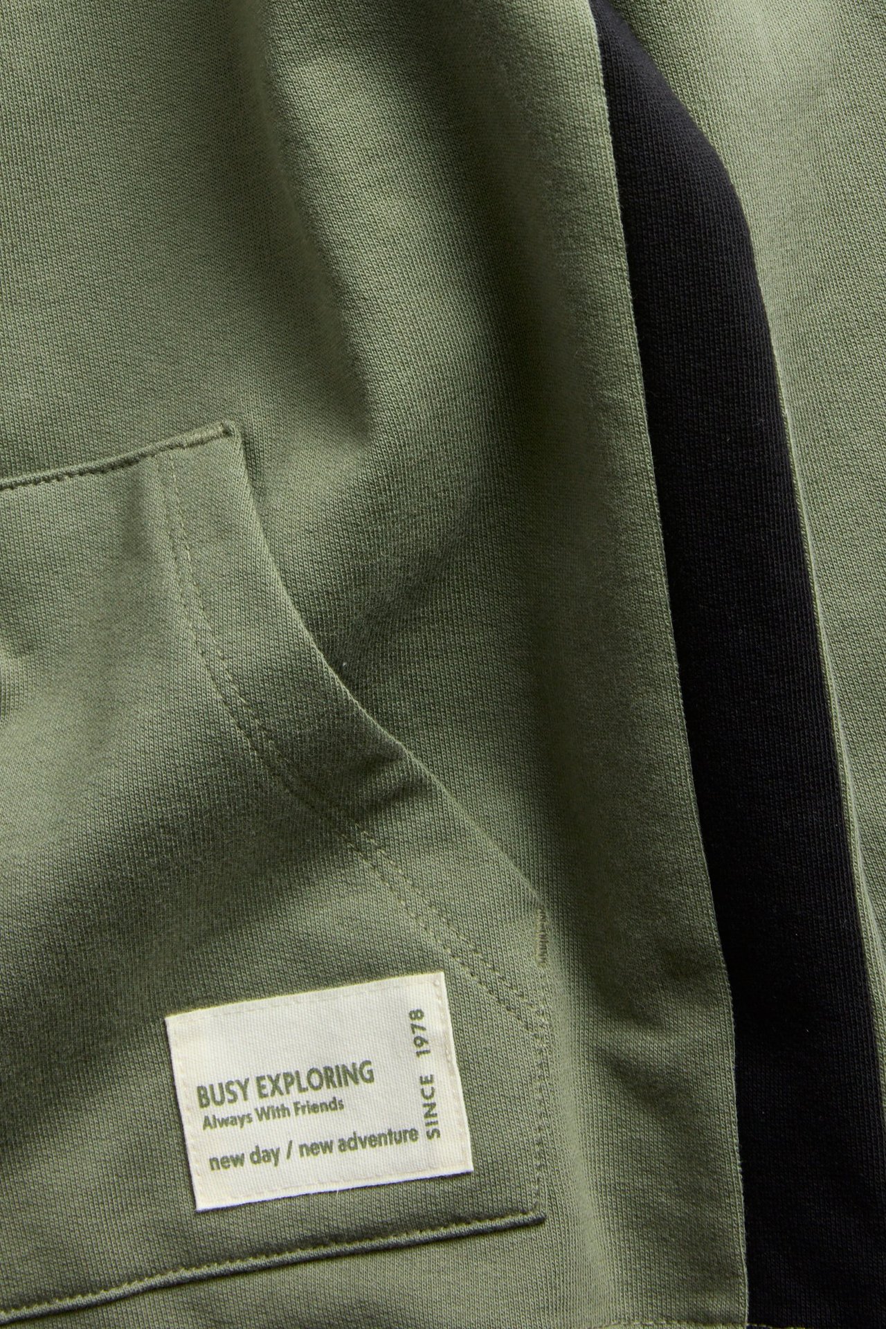 Zbliżenie ciemnozielonej bluzy z kapturem z kieszenią kangurką, białą naszywką i czarnym panelem bocznym.