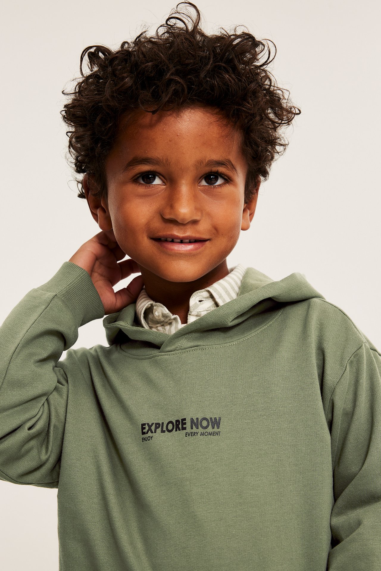 Lähikuva lapsesta yllään tummanvihreä puuvillahuppari tekstillä 'EXPLORE NOW' raidallisen paidan päällä.