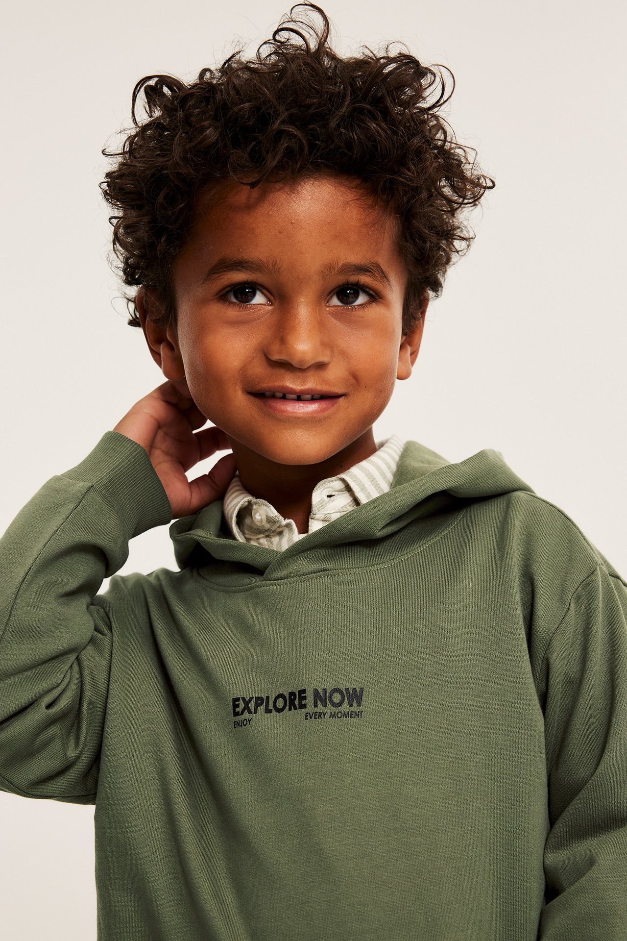 Nærbilde forfra av en smilende gutt i mørkegrønn bomullshoodie over stripet skjorte, hånden ved øret.