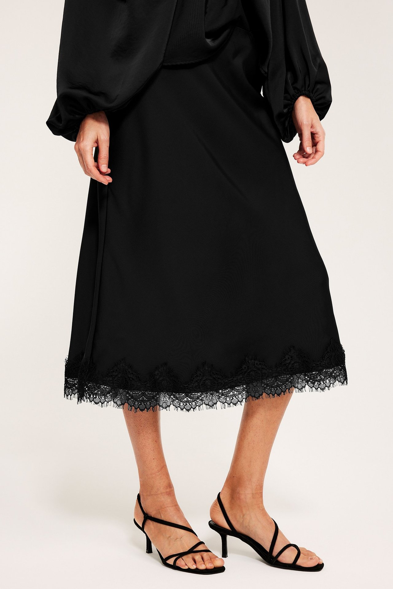 Widok z przodu kobiety w czarnej spódnicy satynowej z koronką, czarnym topie i czarnych sandałach na obcasie.