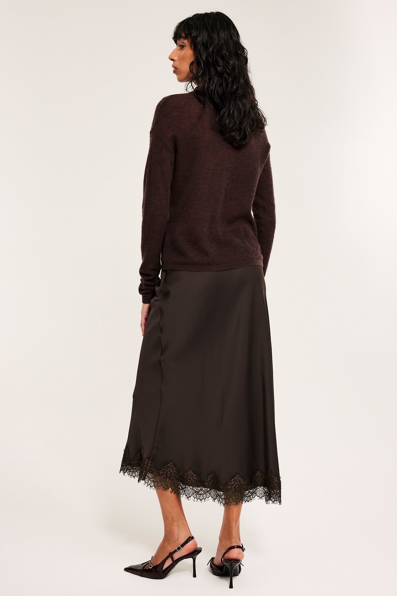 Widok z boku kobiety w brązowej satynowej spódnicy z koronkowym obszyciem, ciemnobrązowym swetrze i szpilkach.