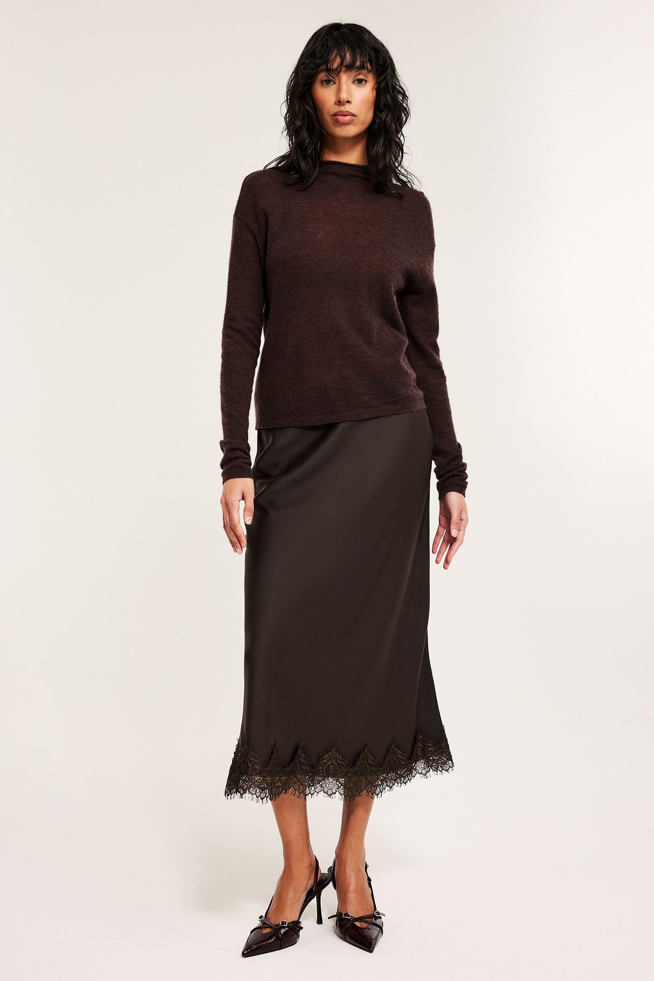 Przodem kobieta w brązowej satynowej spódnicy midi z koronką i ciemnobrązowym swetrze.