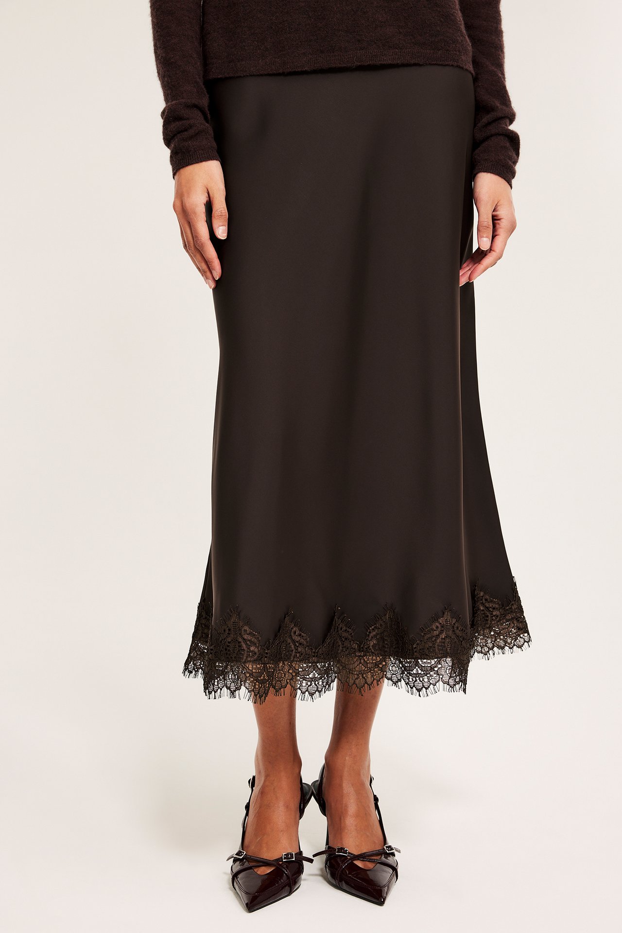 Nainen edestä: ruskea satiinialushame mustalla pitsireunalla, tummanruskea neulepusero ja teräväkärkiset korkokengät.