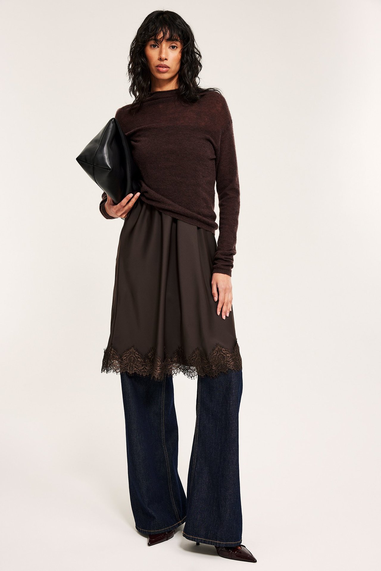 Nainen ruskeassa pitsireunaisessa satiinihameessa, tummanruskea paita, siniset farkut, musta käsilaukku.
